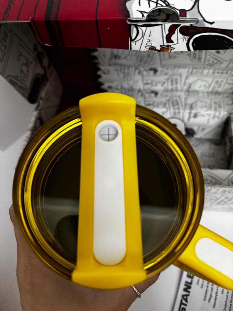 May include: A yellow travel mug with a clear lid and a white sliding closure mechanism. The mug features a yellow handle and is positioned in front of a box decorated with black and white comic strip illustrations of a cartoon dog character.