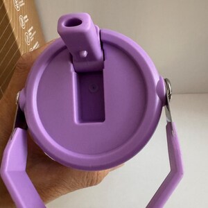 May include: A close-up of a purple water bottle lid with a handle. The lid has a flip-top opening and a circular design. The handle is hexagonal and attached to the lid with silver metal hinges. The lid is held by a hand.
