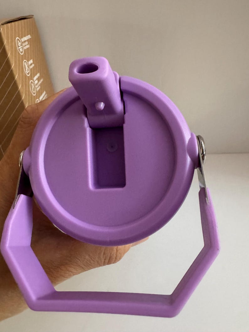 May include: A close-up of a purple water bottle lid with a handle. The lid has a flip-top opening and a circular design. The handle is hexagonal and attached to the lid with silver metal hinges. The lid is held by a hand.