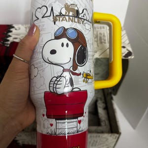 May include: A white Stanley tumbler featuring a graphic of Snoopy wearing aviator goggles and a scarf, sitting on a red doghouse. The cup has a yellow lid and handle, with the text SNOOPY printed in yellow at the base and a comic strip background.
