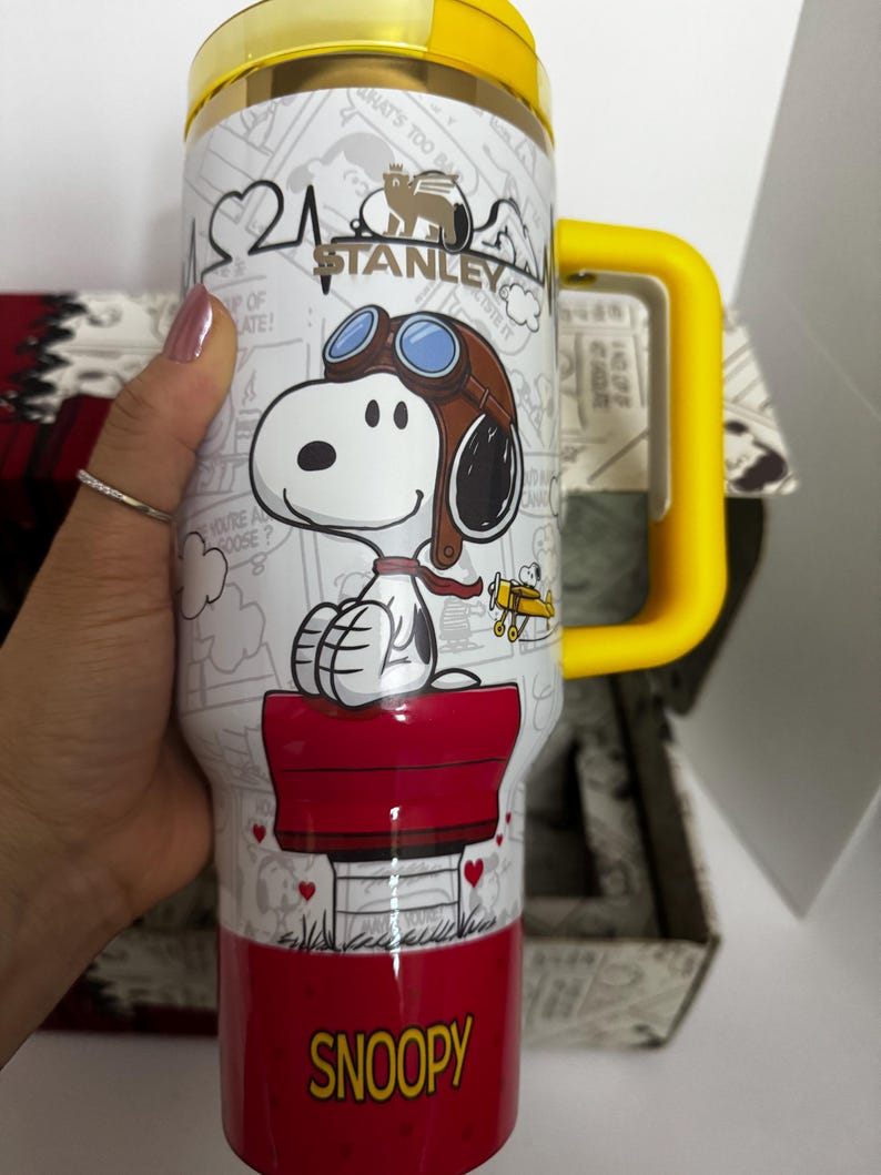 May include: A white Stanley tumbler featuring a graphic of Snoopy wearing aviator goggles and a scarf, sitting on a red doghouse. The cup has a yellow lid and handle, with the text SNOOPY printed in yellow at the base and a comic strip background.