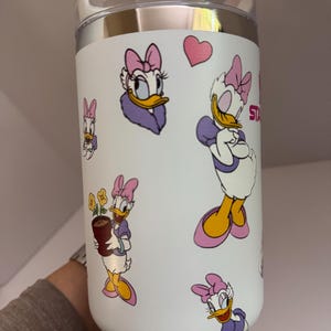Op de afbeelding: Witte ge&iuml;soleerde beker met illustraties van Daisy Duck in verschillende poses. De beker heeft een doorzichtig deksel en een zilveren rand. Daisy Duck is afgebeeld in haar kenmerkende paarse en roze outfit, met een roze hart.