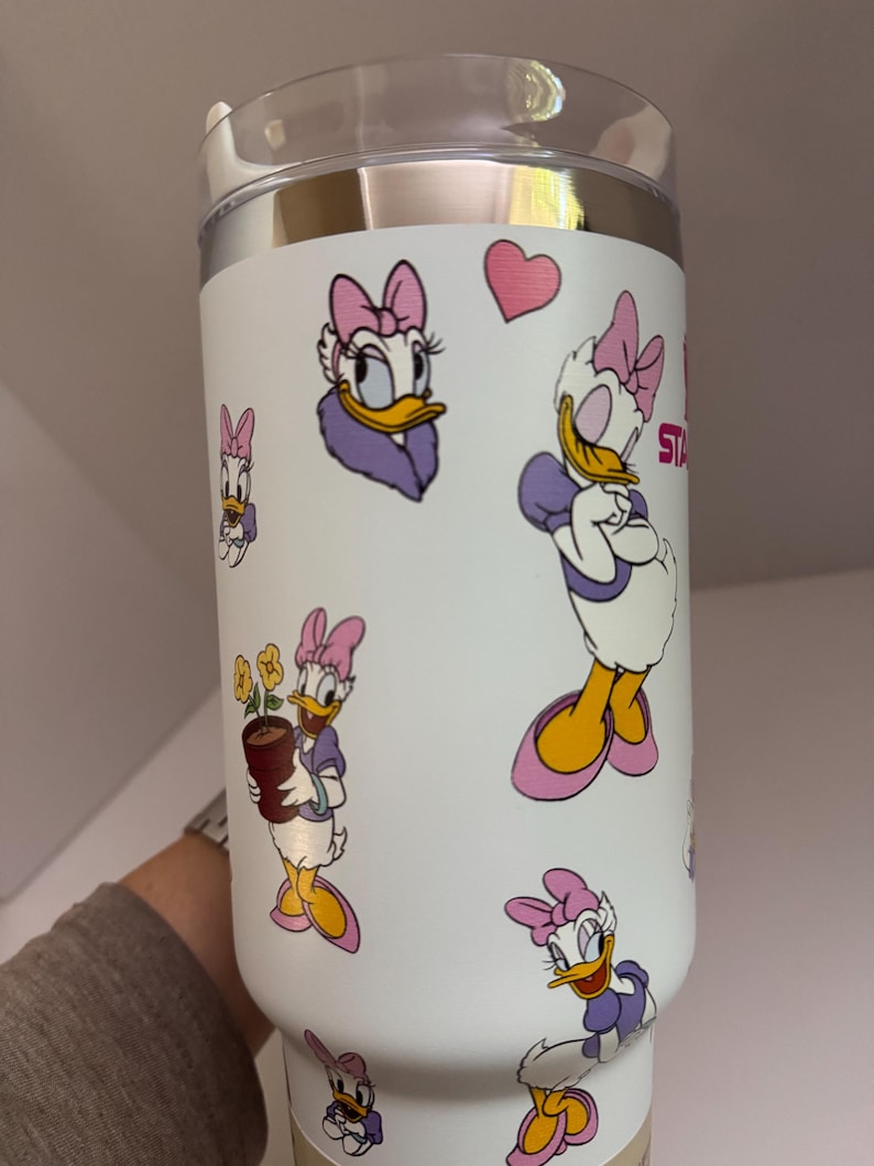 Op de afbeelding: Witte ge&iuml;soleerde beker met illustraties van Daisy Duck in verschillende poses. De beker heeft een doorzichtig deksel en een zilveren rand. Daisy Duck is afgebeeld in haar kenmerkende paarse en roze outfit, met een roze hart.
