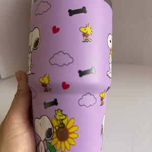 May include: A purple insulated tumbler featuring Snoopy, Woodstock, hearts, clouds, and dog bones. The tumbler has a purple lid and a handle. The design includes Snoopy holding a sunflower. The tumbler is designed to keep drinks cold or hot.