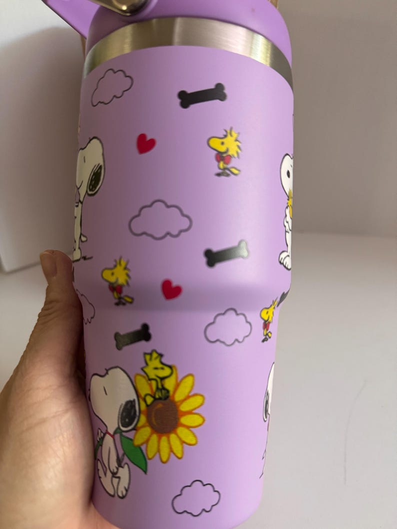 May include: A purple insulated tumbler featuring Snoopy, Woodstock, hearts, clouds, and dog bones. The tumbler has a purple lid and a handle. The design includes Snoopy holding a sunflower. The tumbler is designed to keep drinks cold or hot.