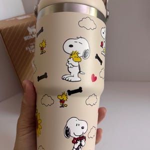 May include: Beige insulated tumbler featuring Snoopy and Woodstock cartoon graphics. The tumbler has a cream-colored lid and handle, with a stainless steel band. The design includes cartoon clouds, hearts, and bones. The tumbler is designed to keep beverages cold or hot.