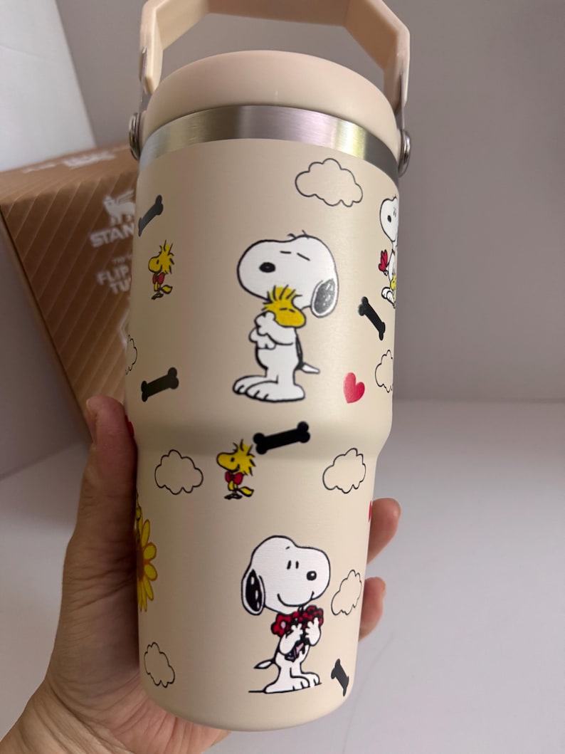 May include: Beige insulated tumbler featuring Snoopy and Woodstock cartoon graphics. The tumbler has a cream-colored lid and handle, with a stainless steel band. The design includes cartoon clouds, hearts, and bones. The tumbler is designed to keep beverages cold or hot.