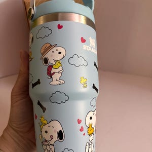 May include: A light blue Stanley tumbler featuring Snoopy and Woodstock cartoon graphics. The stainless steel tumbler has a light blue handle and lid, with a silver band at the top. The design includes hearts, clouds, and bone shapes.