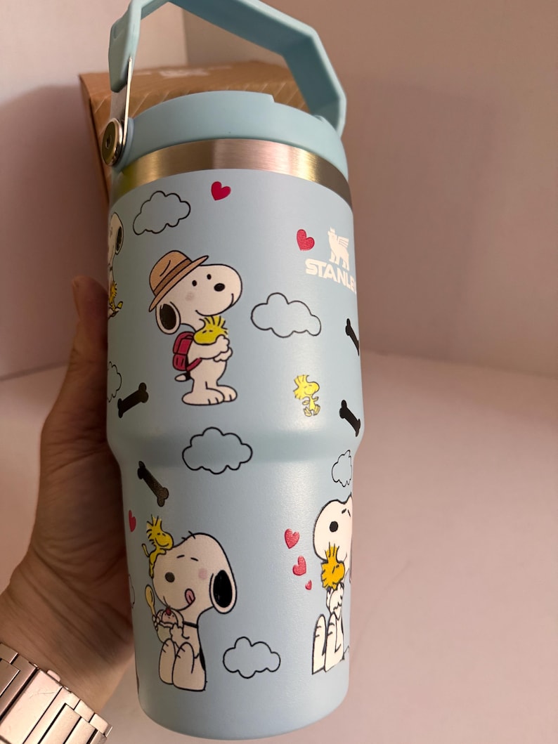 May include: A light blue Stanley tumbler featuring Snoopy and Woodstock cartoon graphics. The stainless steel tumbler has a light blue handle and lid, with a silver band at the top. The design includes hearts, clouds, and bone shapes.