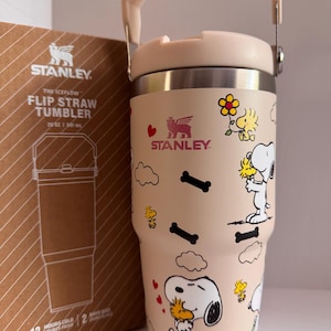 May include: A beige Stanley The Iceflow Flip Straw Tumbler, 20 oz, featuring Snoopy and Woodstock cartoon graphics. The tumbler has a light pink handle and lid, with the Stanley logo. The box next to it shows the tumbler's outline and the text "12 HOURS COLD".
