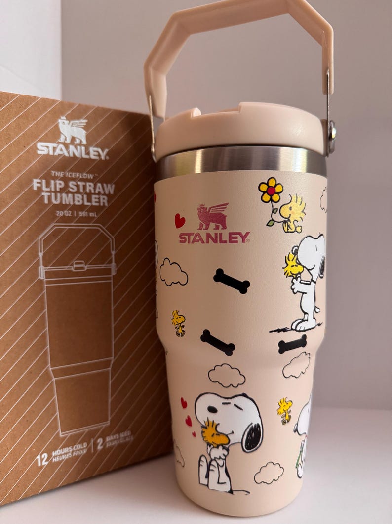 May include: A beige Stanley The Iceflow Flip Straw Tumbler, 20 oz, featuring Snoopy and Woodstock cartoon graphics. The tumbler has a light pink handle and lid, with the Stanley logo. The box next to it shows the tumbler's outline and the text "12 HOURS COLD".