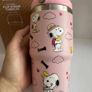 May include: Pink Stanley Flip Straw Tumbler with Snoopy and Woodstock cartoon graphics. The tumbler has a pink handle and lid, and the text "THE ICEFLOW" is visible on the box. The tumbler holds 20 oz.