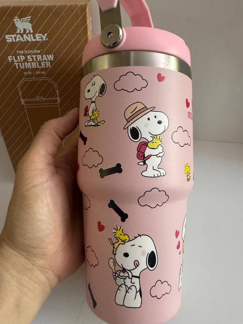 May include: Pink Stanley Flip Straw Tumbler with Snoopy and Woodstock cartoon graphics. The tumbler has a pink handle and lid, and the text "THE ICEFLOW" is visible on the box. The tumbler holds 20 oz.