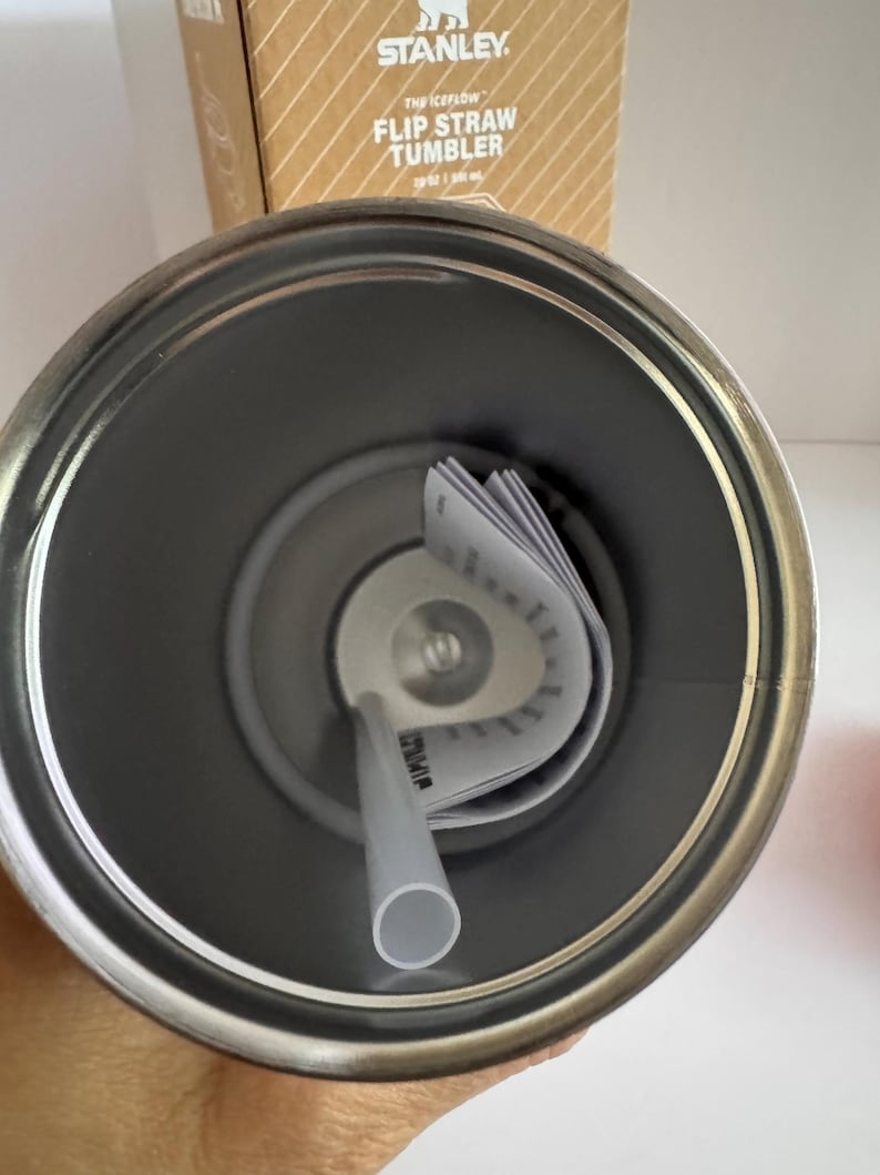 May include: A stainless steel Stanley Flip Straw Tumbler, 30 oz capacity, with a gray straw and interior. The tumbler's interior is visible, along with the straw and a folded product information tag. The top of the tumbler is open, revealing the inside.