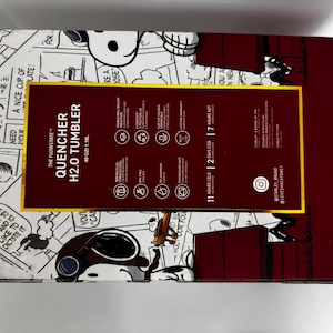 May include: A rectangular product box for a 40 oz Stanley Quencher H2.0 tumbler featuring a Peanuts comic strip design. The box has a dark red central panel with white text detailing product features, including 11 hours cold and 2 days iced.