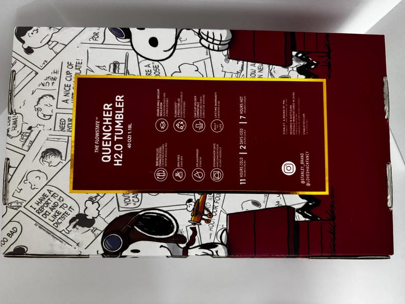 May include: A rectangular product box for a 40 oz Stanley Quencher H2.0 tumbler featuring a Peanuts comic strip design. The box has a dark red central panel with white text detailing product features, including 11 hours cold and 2 days iced.