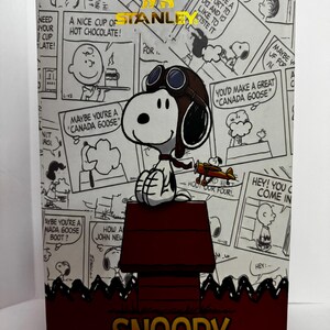 May include: A rectangular product box featuring a black and white Peanuts comic strip background. The box displays a central illustration of Snoopy wearing aviator goggles on a red doghouse, with a gold Stanley logo and the name SNOOPY printed below.