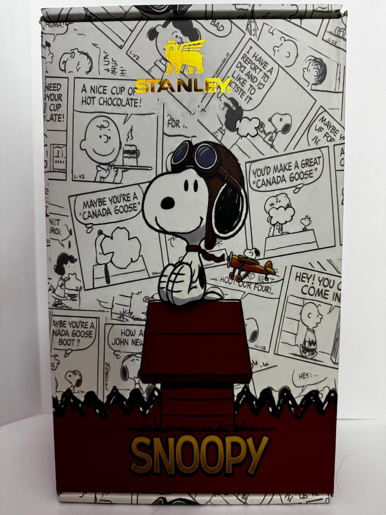 May include: A rectangular product box featuring a black and white Peanuts comic strip background. The box displays a central illustration of Snoopy wearing aviator goggles on a red doghouse, with a gold Stanley logo and the name SNOOPY printed below.