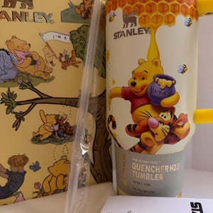 May include: A yellow Stanley Quencher H2.0 tumbler featuring Winnie the Pooh and Tigger illustrations. The 40 oz (1.18L) tumbler has a honey-themed design with a handle and a straw. The packaging includes Winnie the Pooh illustrations.