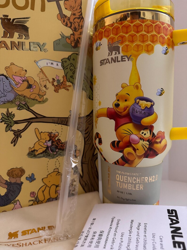 May include: A yellow Stanley Quencher H2.0 tumbler featuring Winnie the Pooh and Tigger illustrations. The 40 oz (1.18L) tumbler has a honey-themed design with a handle and a straw. The packaging includes Winnie the Pooh illustrations.