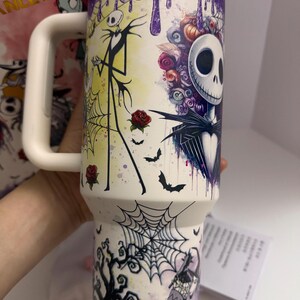 Puede incluir: Un vaso aislado blanco con un dise&ntilde;o colorido de Jack Skellington de The Nightmare Before Christmas. El dise&ntilde;o incluye murci&eacute;lagos, rosas y una telara&ntilde;a. La parte superior tiene un dise&ntilde;o de goteo p&uacute;rpura y un asa blanca.