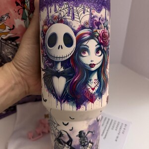 Puede incluir: Un vaso blanco con un dise&ntilde;o que presenta a Jack Skellington y Sally de The Nightmare Before Christmas. El dise&ntilde;o incluye un efecto de goteo p&uacute;rpura en la parte superior y una escena con otros personajes en la parte inferior. El vaso es sostenido por una mano.