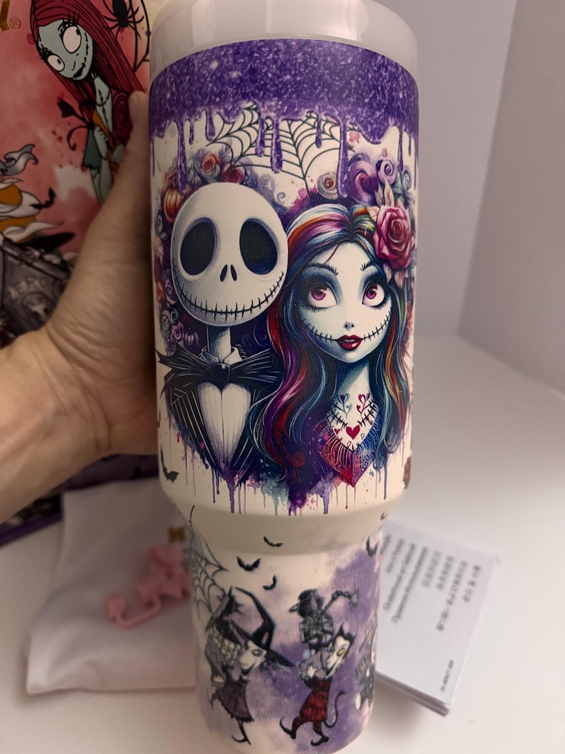 Puede incluir: Un vaso blanco con un dise&ntilde;o que presenta a Jack Skellington y Sally de The Nightmare Before Christmas. El dise&ntilde;o incluye un efecto de goteo p&uacute;rpura en la parte superior y una escena con otros personajes en la parte inferior. El vaso es sostenido por una mano.
