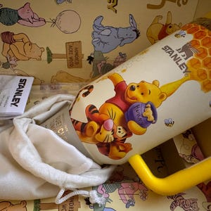 May include: A Stanley brand Winnie the Pooh themed tumbler with a yellow handle. The tumbler features a cartoon illustration of Winnie the Pooh and Tigger with a honey pot. The tumbler is in a box with a Winnie the Pooh theme.
