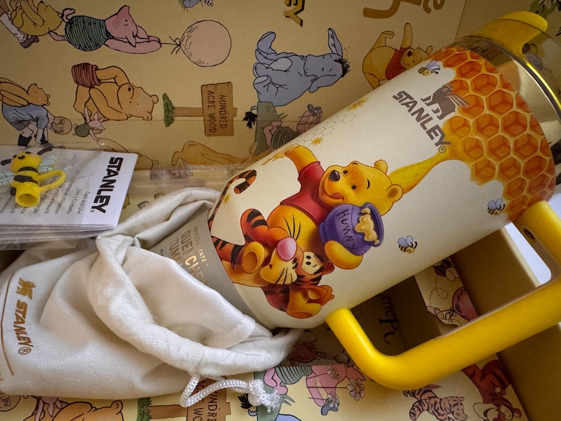 May include: A Stanley brand Winnie the Pooh themed tumbler with a yellow handle. The tumbler features a cartoon illustration of Winnie the Pooh and Tigger with a honey pot. The tumbler is in a box with a Winnie the Pooh theme.
