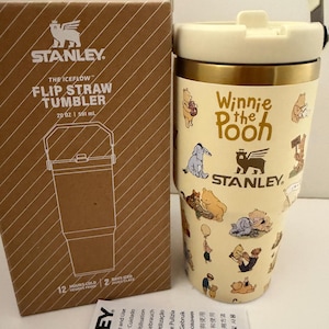 Puede incluir: Un vaso Stanley IceFlow de color crema con pajita abatible y capacidad de 591 ml, decorado con ilustraciones de Winnie the Pooh, borde dorado y tapa a juego con asa, mostrado junto a su embalaje de cartón de color marrón.