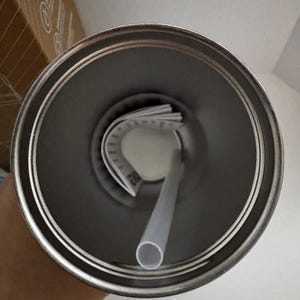 Puede incluir: Un vaso de acero inoxidable con un borde plateado, una pajita de pl&aacute;stico transparente y una etiqueta blanca doblada en el interior. El vaso est&aacute; vac&iacute;o y se ve desde arriba, mostrando el interior.