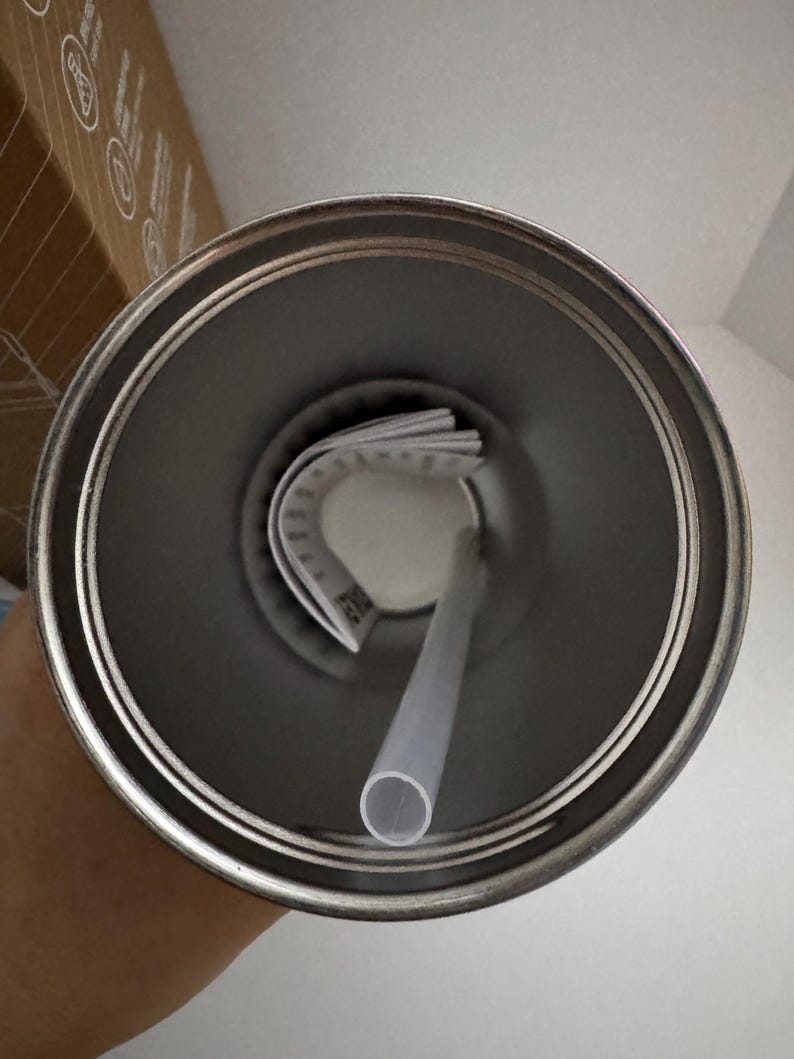 Puede incluir: Un vaso de acero inoxidable con un borde plateado, una pajita de pl&aacute;stico transparente y una etiqueta blanca doblada en el interior. El vaso est&aacute; vac&iacute;o y se ve desde arriba, mostrando el interior.