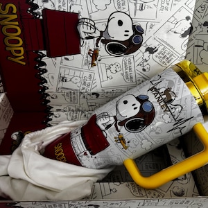 May include: A white Stanley tumbler featuring a Snoopy graphic with a pilot hat and goggles, accented by a yellow handle and lid. The tumbler is presented in a matching box decorated with black and white Peanuts comic strip panels and Snoopy text.