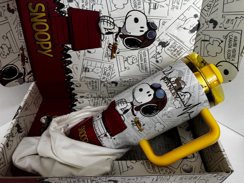 May include: A white Stanley tumbler featuring a Snoopy graphic with a pilot hat and goggles, accented by a yellow handle and lid. The tumbler is presented in a matching box decorated with black and white Peanuts comic strip panels and Snoopy text.
