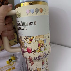 K&ouml;nnte beinhalten: Ein beigefarbener und goldfarbener Isolierbecher mit Winnie-Puuh-Figuren und Luftballons. Der Becher hat einen Griff und einen hellblauen Deckel. Der Text "QUENCHER H2.0 TUMBLER" ist auf der Seite zu sehen.