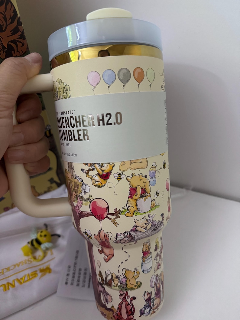 K&ouml;nnte beinhalten: Ein beigefarbener und goldfarbener Isolierbecher mit Winnie-Puuh-Figuren und Luftballons. Der Becher hat einen Griff und einen hellblauen Deckel. Der Text "QUENCHER H2.0 TUMBLER" ist auf der Seite zu sehen.