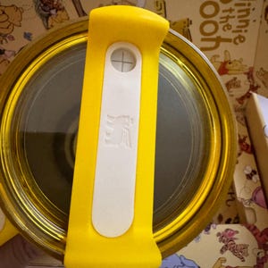 May include: A yellow and gold Winnie the Pooh themed container with a yellow handle and a white button. The container has a clear, reflective surface. The handle has a white section with a cross symbol and a logo.