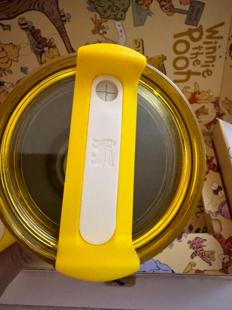 May include: A yellow and gold Winnie the Pooh themed container with a yellow handle and a white button. The container has a clear, reflective surface. The handle has a white section with a cross symbol and a logo.