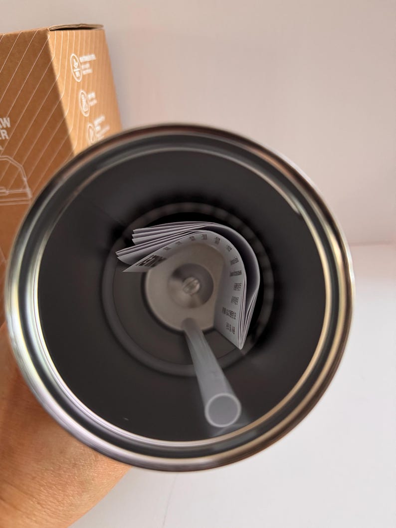 May include: A stainless steel tumbler with a gray straw and a paper insert. The interior of the tumbler is dark gray, and the straw is light gray. The tumbler is designed to keep drinks cold or hot.