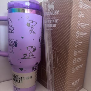 Może przedstawiać: Fioletowy kubek Stanley Quencher H2.0 Flowstate z wzorami Snoopy'ego i Woodstocka. Kubek ma uchwyt, pokrywkę i słomkę. Pudełko jest brązowe z przekątnymi liniami i logo Stanley.
