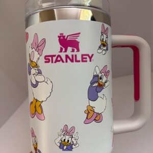 Op de afbeelding: Witte Stanley reismok met Daisy Duck illustraties. De mok heeft een roze handvat en accenten, een doorzichtig deksel en het Stanley logo. Het ontwerp toont Daisy Duck in paarse en roze outfits.
