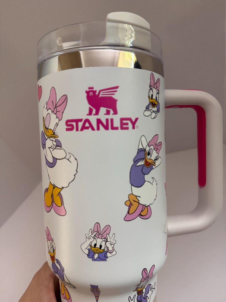 Op de afbeelding: Witte Stanley reismok met Daisy Duck illustraties. De mok heeft een roze handvat en accenten, een doorzichtig deksel en het Stanley logo. Het ontwerp toont Daisy Duck in paarse en roze outfits.