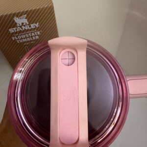 May include: A pink Stanley Flowstate tumbler lid with a matching handle. The lid has a sliding closure and a clear, transparent rim. The box in the background has the Stanley logo and the words "Flowstate Tumbler".