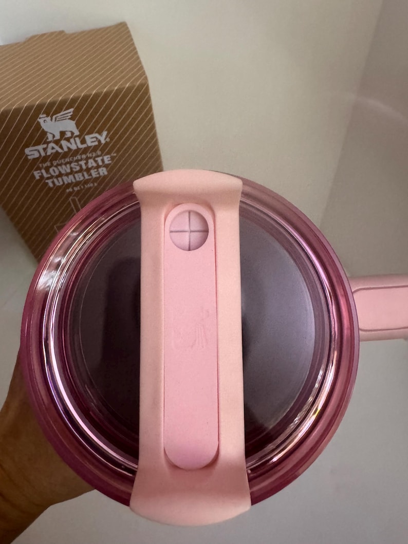 May include: A pink Stanley Flowstate tumbler lid with a matching handle. The lid has a sliding closure and a clear, transparent rim. The box in the background has the Stanley logo and the words "Flowstate Tumbler".