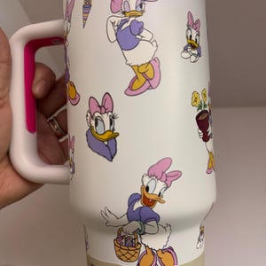 Op de afbeelding: Witte ge&iuml;soleerde beker met Daisy Duck cartoonafbeeldingen in paars, roze en geel. De beker heeft een witte handgreep met een roze accent. Houdt drankjes tot 2 dagen koud.