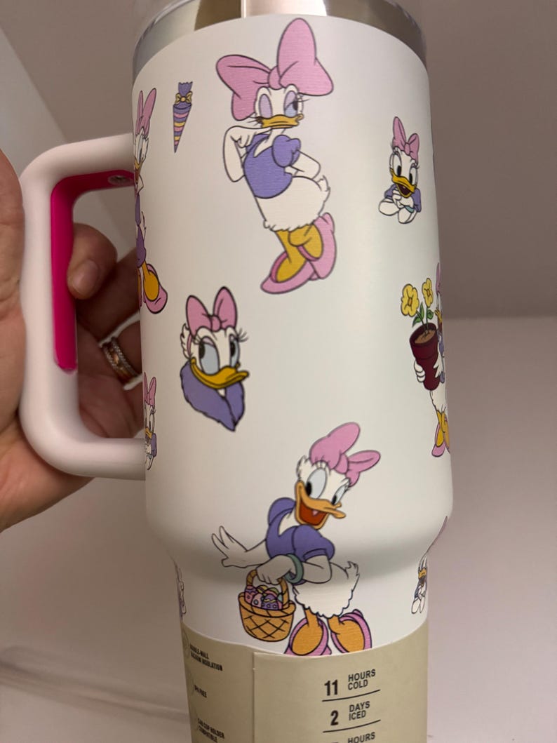 Op de afbeelding: Witte ge&iuml;soleerde beker met Daisy Duck cartoonafbeeldingen in paars, roze en geel. De beker heeft een witte handgreep met een roze accent. Houdt drankjes tot 2 dagen koud.