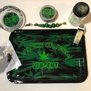 May include: A green and black rolling tray with a marijuana leaf design and the text "It's a Romero thing... Fuckery! Vincent". The tray is surrounded by a variety of smoking accessories, including a glass ashtray, a small glass jar, a lighter, and a beaded necklace.
