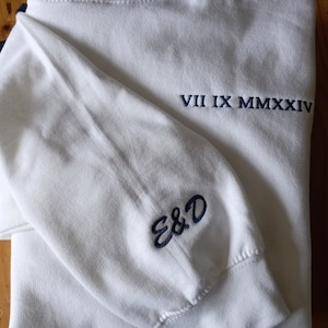 May include: White long-sleeved sweatshirt with navy blue embroidered text "VII IX MMXXXIV" on the back and "E&D" on the sleeve.