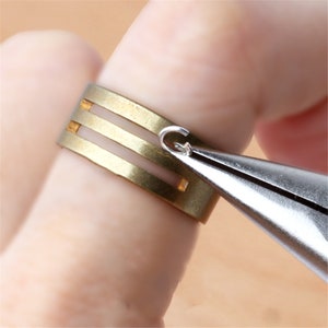 May include: A gold ring with three horizontal lines is being attached to a silver jump ring with a pair of pliers.