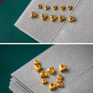 May include: A collection of small, gold-colored triangle-shaped beads with a hole in the center. The beads are arranged in a pattern on a gray surface.